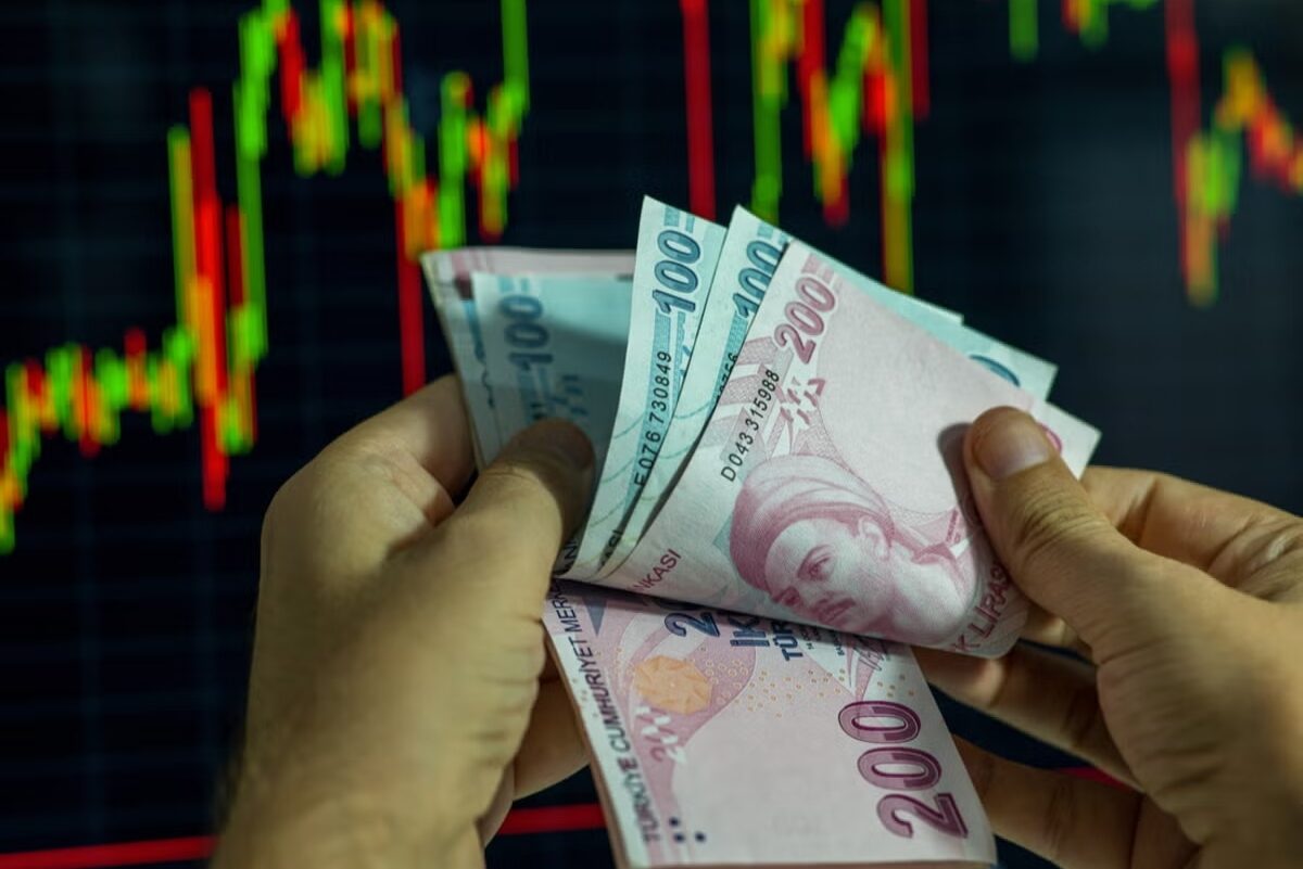 Hands holding Turkish lira in front of a financial screen.